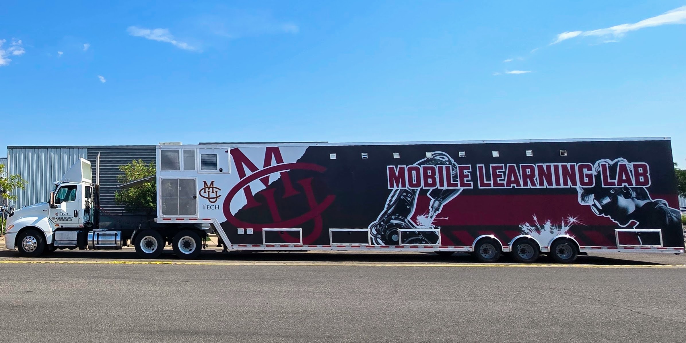 The image shows a large CMU Tech Mobile Learning Lab trailer attached to a white semi-truck parked along a street under a clear blue sky. The trailer is painted in black, maroon, and white with bold text reading “Mobile Learning Lab.” On the left side of the trailer, the CMU Tech logo is displayed prominently. The design also features a large robotic arm illustration and a profile image of a person wearing protective welding goggles, with sparks shown to represent welding. The trailer has three axles and multiple access panels along the lower side.